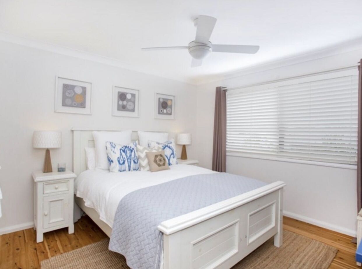 A bedroom with a white bed, two bedside tables, a ceiling fan, and a window with blinds.