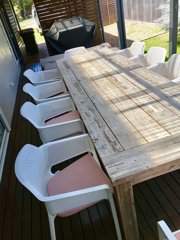Um deck de madeira com uma mesa grande e cadeiras de plástico branco.