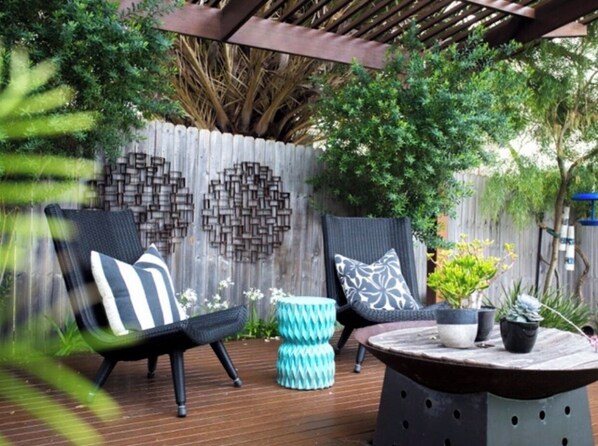 Relax in a serene outdoor space with a wooden deck, two comfortable chairs, and a round table surrounded by lush greenery.