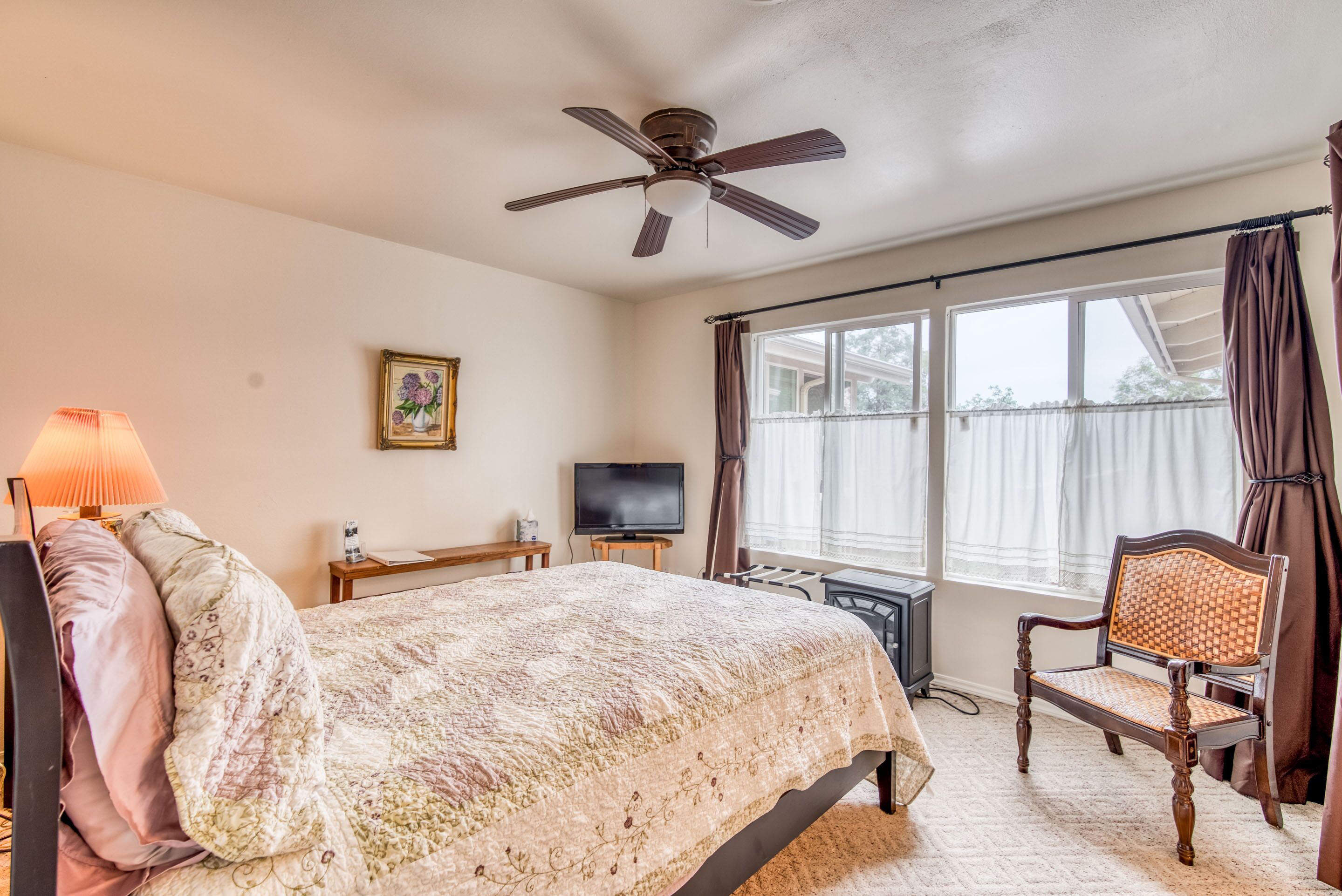 Un dormitorio con cama, ventilador de techo, televisor, una silla y ventana con cortinas.