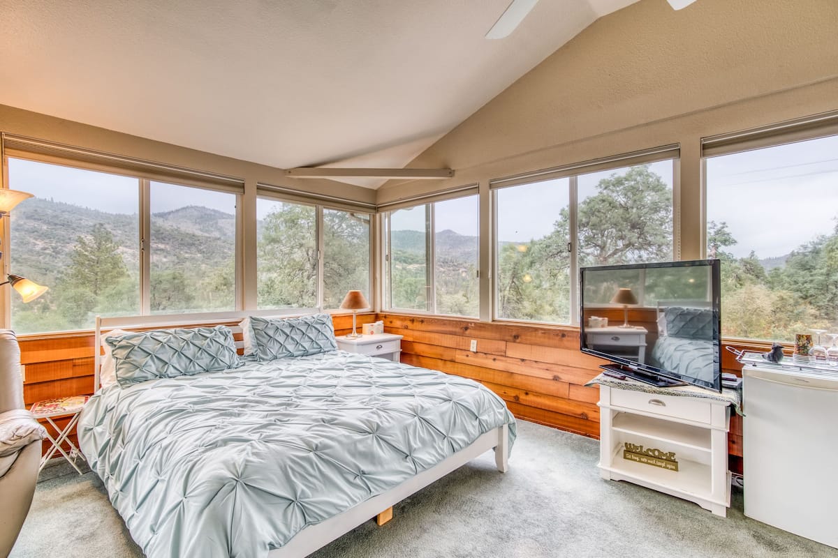Ein Schlafzimmer mit Bett, Fernseher, Schreibtisch und großen Fenstern mit Blick ins Freie.