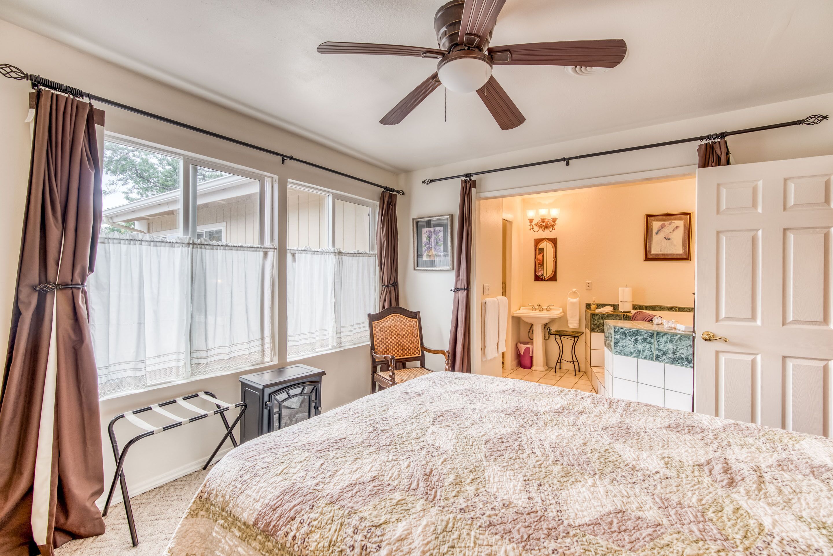 Quarto com cama, ventoinha no teto, cadeira, janela com cortinas e banheiro visível por uma porta aberta.