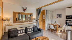 A cozy living room with a sofa, coffee table, and a dining area with a table and chairs. A large mirror hangs above the sofa, and a deer head decoration is on the wall. - Apartment Cry - Alpes Agency - Central Chamonix - Sleeps 2-4 (Chamonix-Mont-Blanc)