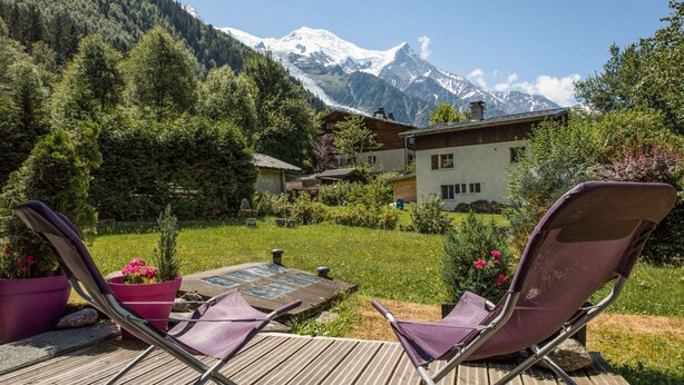 A wooden deck with two purple lounge chairs, a stone pathway, and a house with a mountain backdrop. - Apartment Cry - Alpes Agency - Central Chamonix - Sleeps 2-4 (Chamonix-Mont-Blanc)