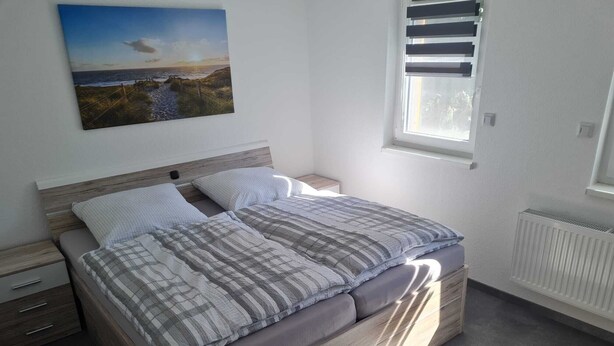 A bedroom with a bed, a nightstand, a drawer unit, a window with blinds, and a wall-mounted picture. - Ferienwohnung 9 & 40 (Stedesdorf)