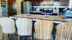 Una cocina con armarios de madera oscura, un refrigerador de acero inoxidable, salpicadero de azulejos y una isla central con cuatro sillas tejidas.
