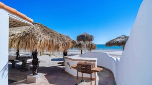 A beachside patio with thatched umbrellas, a barbecue grill, and a stone bench. - La Rosa del Encanto Beachfront Ground Floor West (Playa Encanto)