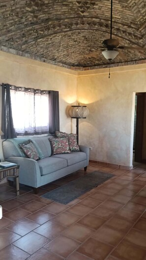 A room with a stone ceiling, a sofa with patterned cushions, a floor lamp, and a window with curtains.
