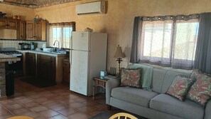 Cocina compacta con refrigerador, estufa y lavamanos. Hay un sofá con cojines decorativos y una lámpara.