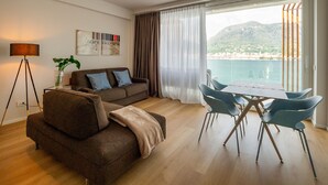 A modern living room with a sofa, a dining table with chairs, a floor lamp, and a view of the sea through large windows. - Ah Porticcioli boutique apartments (San Felice del Benaco)