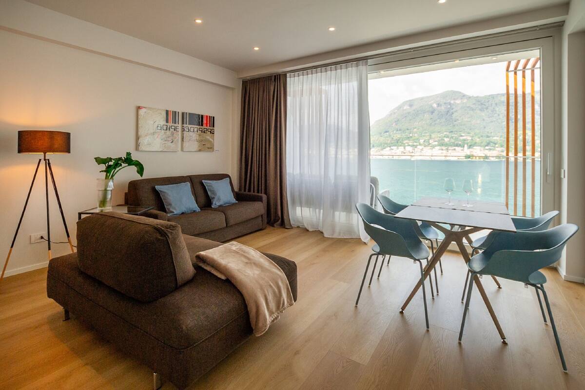 A modern living room with a sofa, a dining table with chairs, a floor lamp, and a view of the sea through large windows.