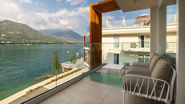 A balcony with a view of a lake and mountains, a white chair, and a glass railing. - Ah Porticcioli boutique apartments (San Felice del Benaco)