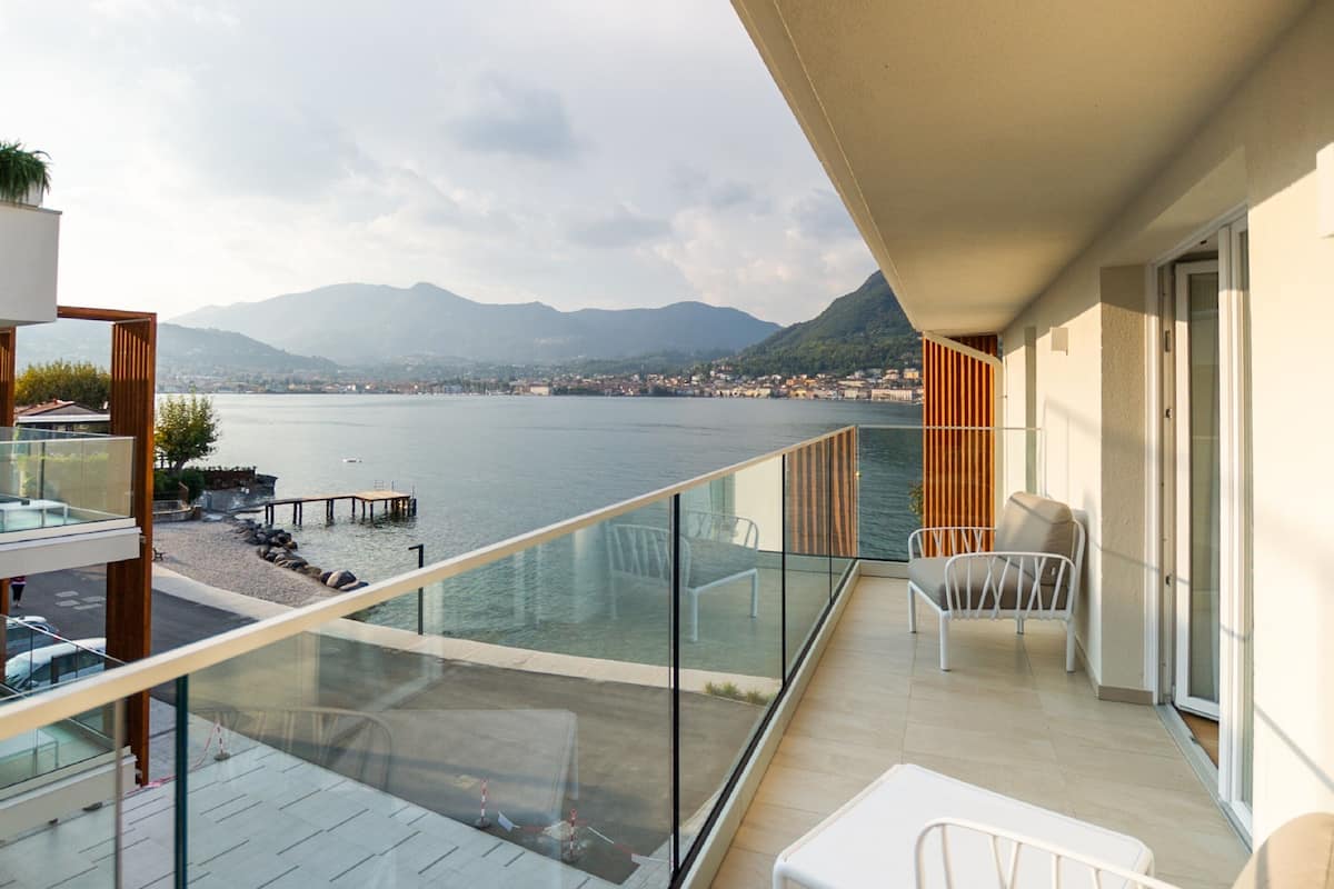 A balcony with a glass railing overlooking a lake and mountains. There are white chairs and a table on the balcony.