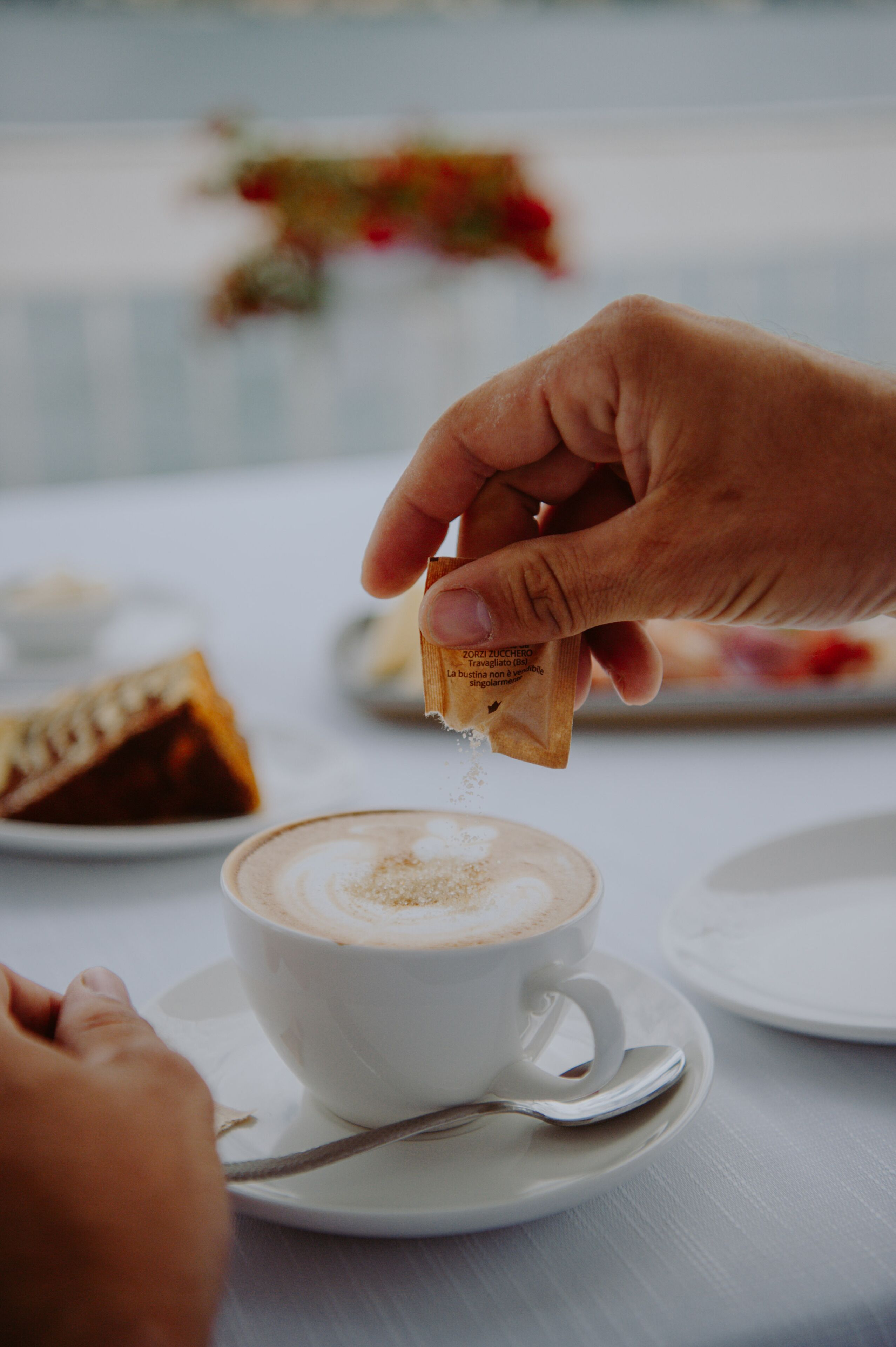 Una mano espolvoreando azúcar en una taza de café sobre una mesa blanca.