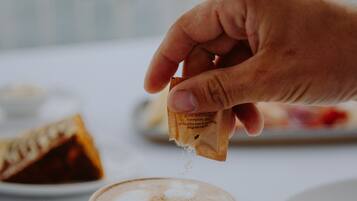 Eine Hand streut Zucker in eine Kaffeetasse auf einem weißen Tisch.
