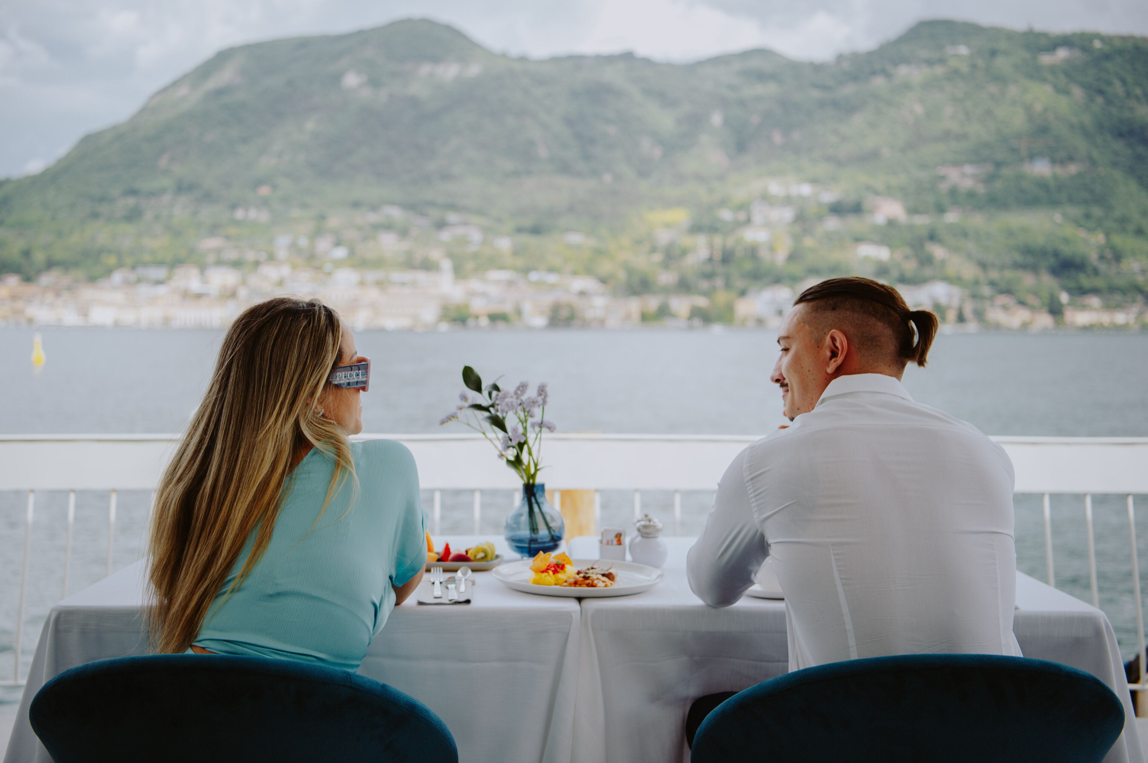 A couple dining at a table with a view of a mountainous landscape and a body of water.