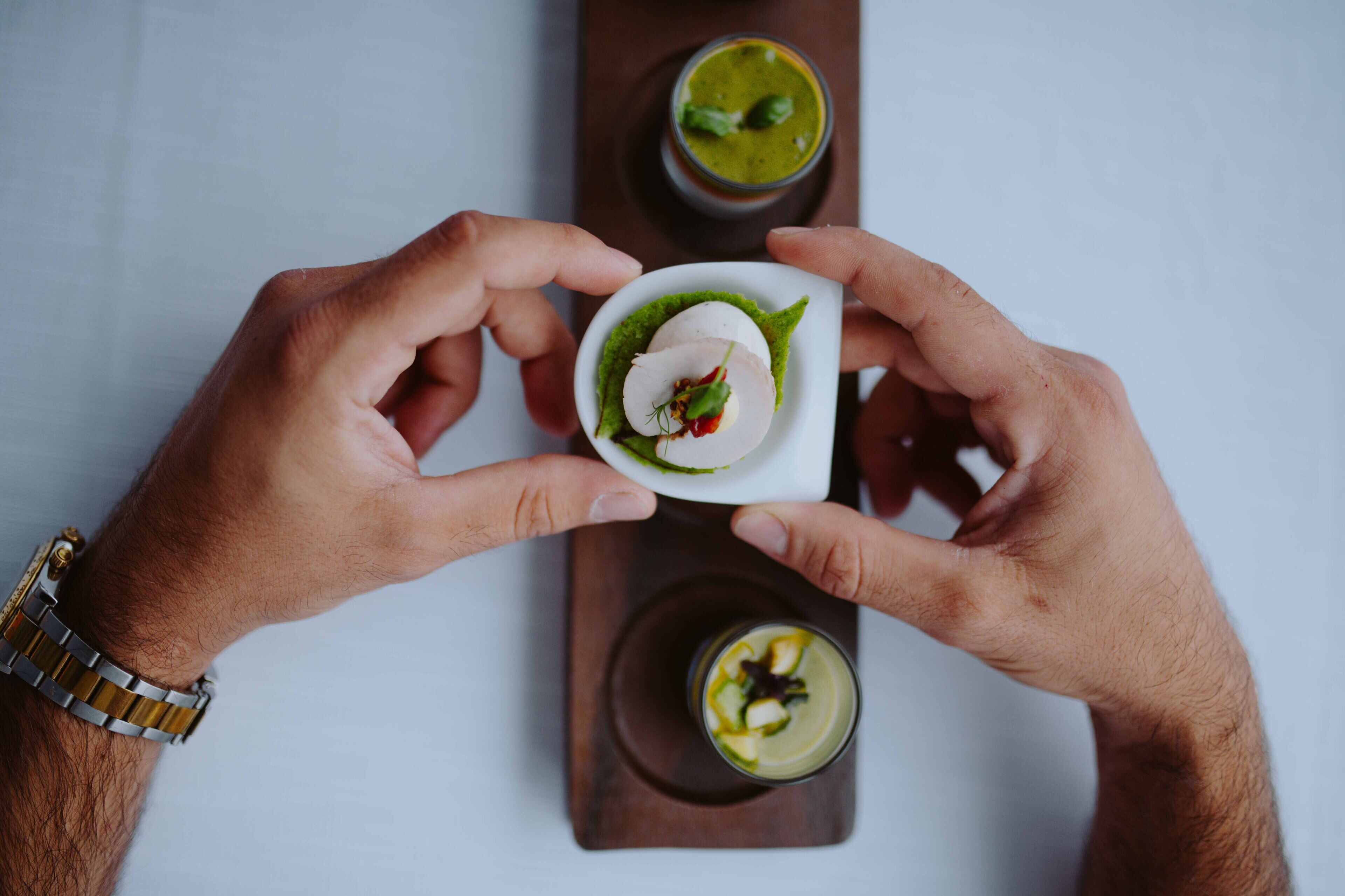 Iemand houdt een klein bord vast met een groene saus en een witte inhoud, mogelijk een dessert, met een houten plank en een glas groene saus op de achtergrond.