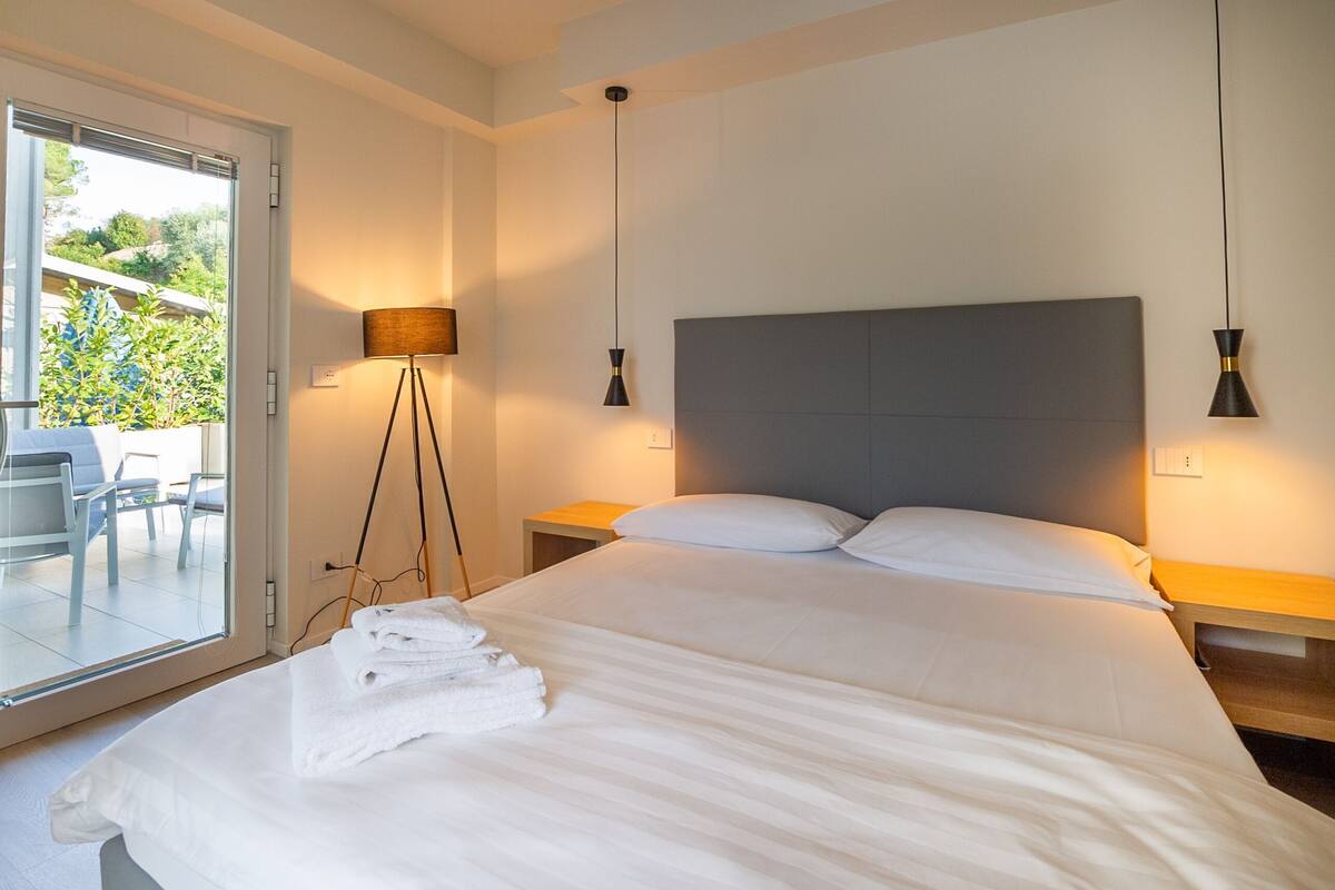 A bedroom with a large bed, bedside lamps, a floor lamp, a glass door leading to an outdoor area, and a small table with a chair.