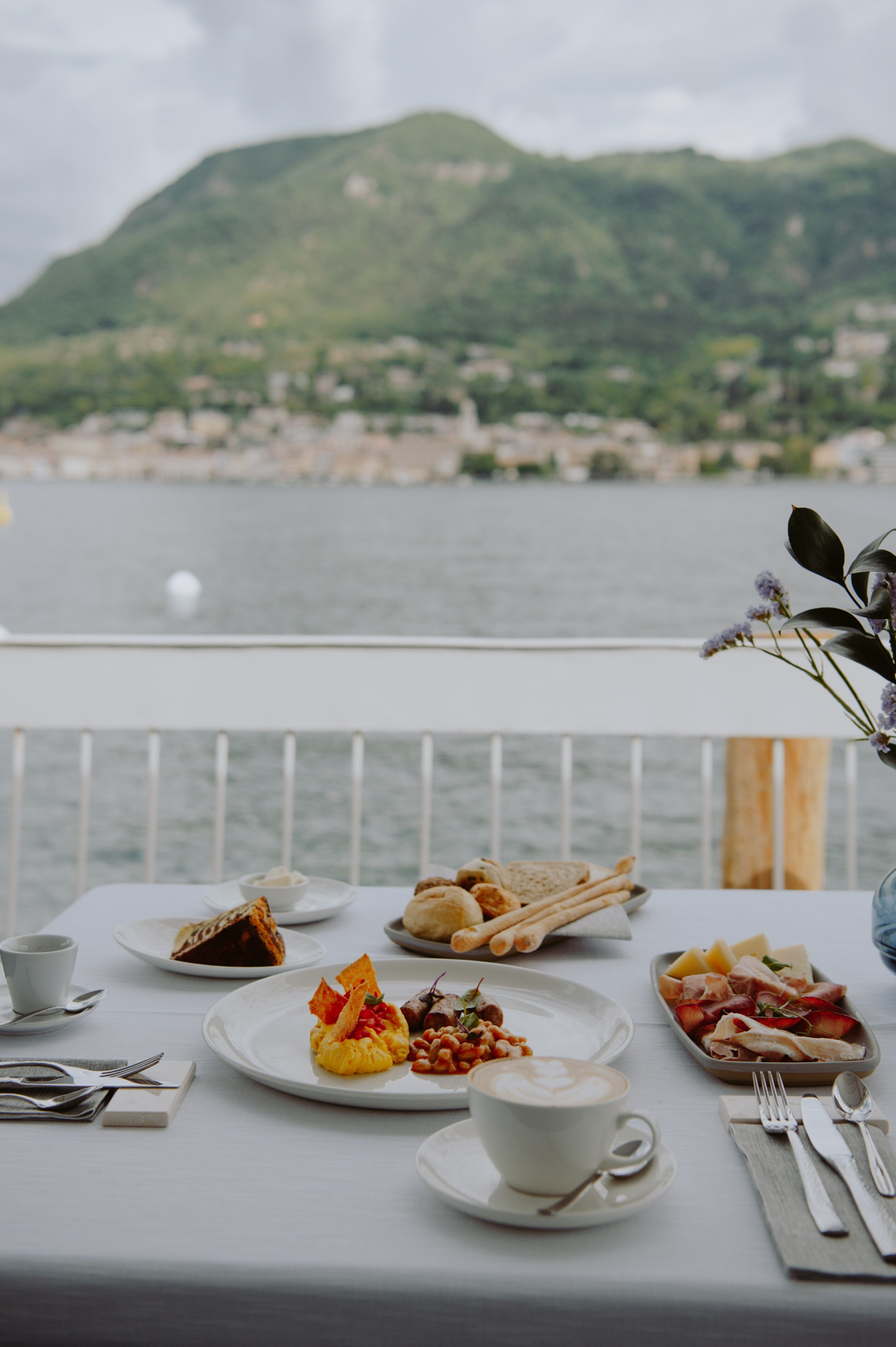 A dining table with various dishes and a cup of coffee, overlooking a body of water and mountains.