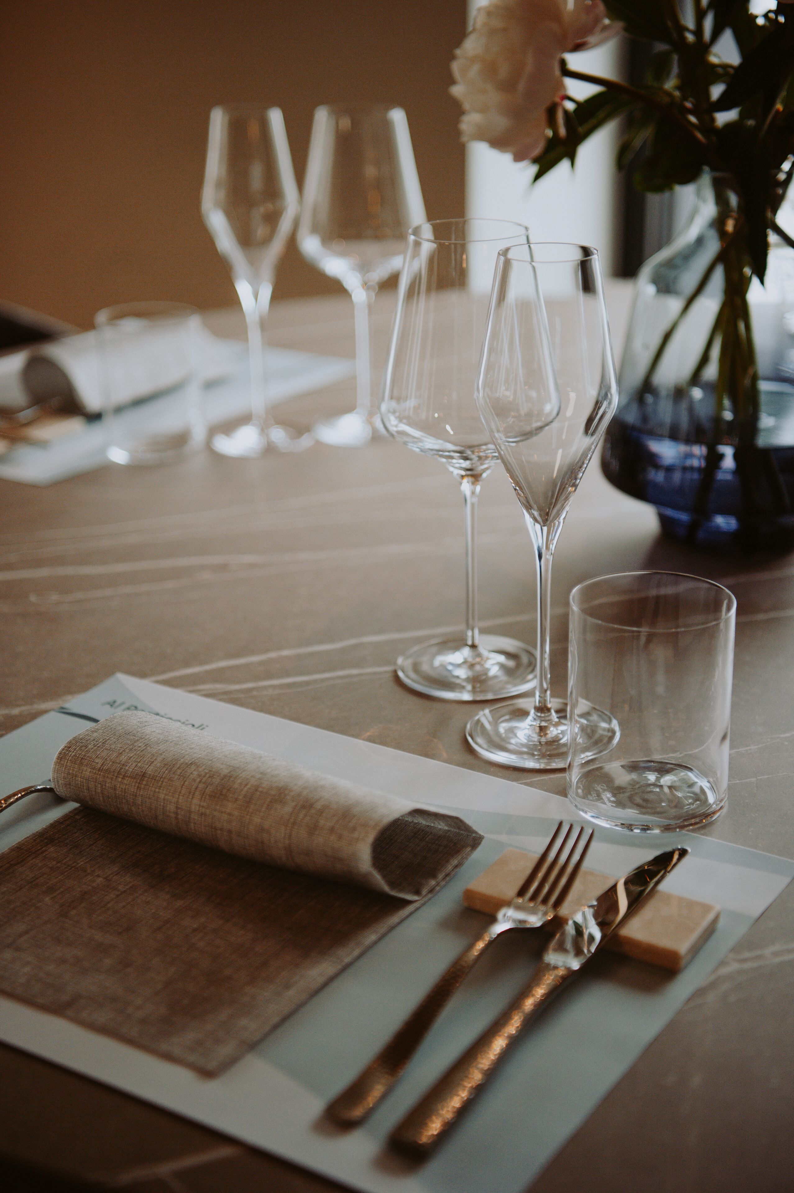 Una mesa dispuesta con servilleta, cubiertos y copas de vino, con un florero en el fondo.