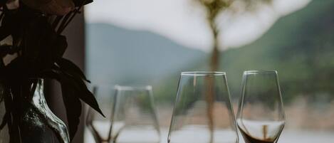 Una mesa con tres copas de vino y un florero con flores, frente a un árbol y montañas.