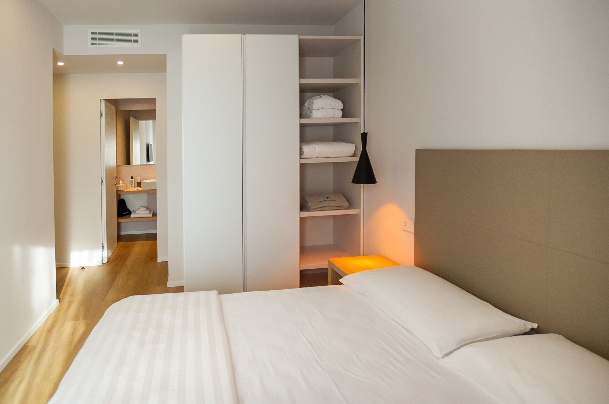 A hotel room with a bed, a wardrobe, a shelf with folded towels, a bedside table with a lamp, and a bathroom visible through an open door.