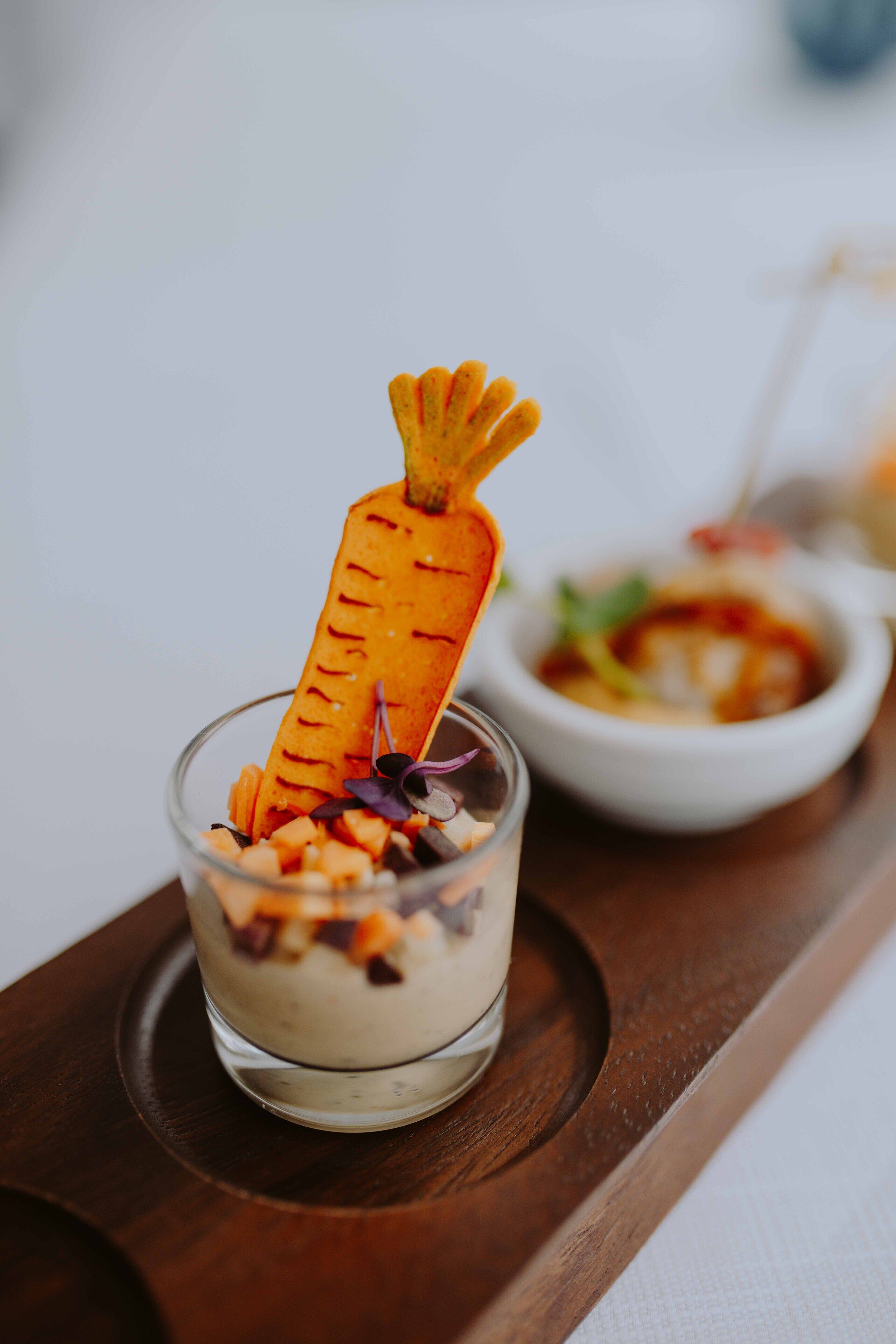 A dessert with a carrot-shaped garnish on a glass dish, accompanied by a small bowl of food in the background.