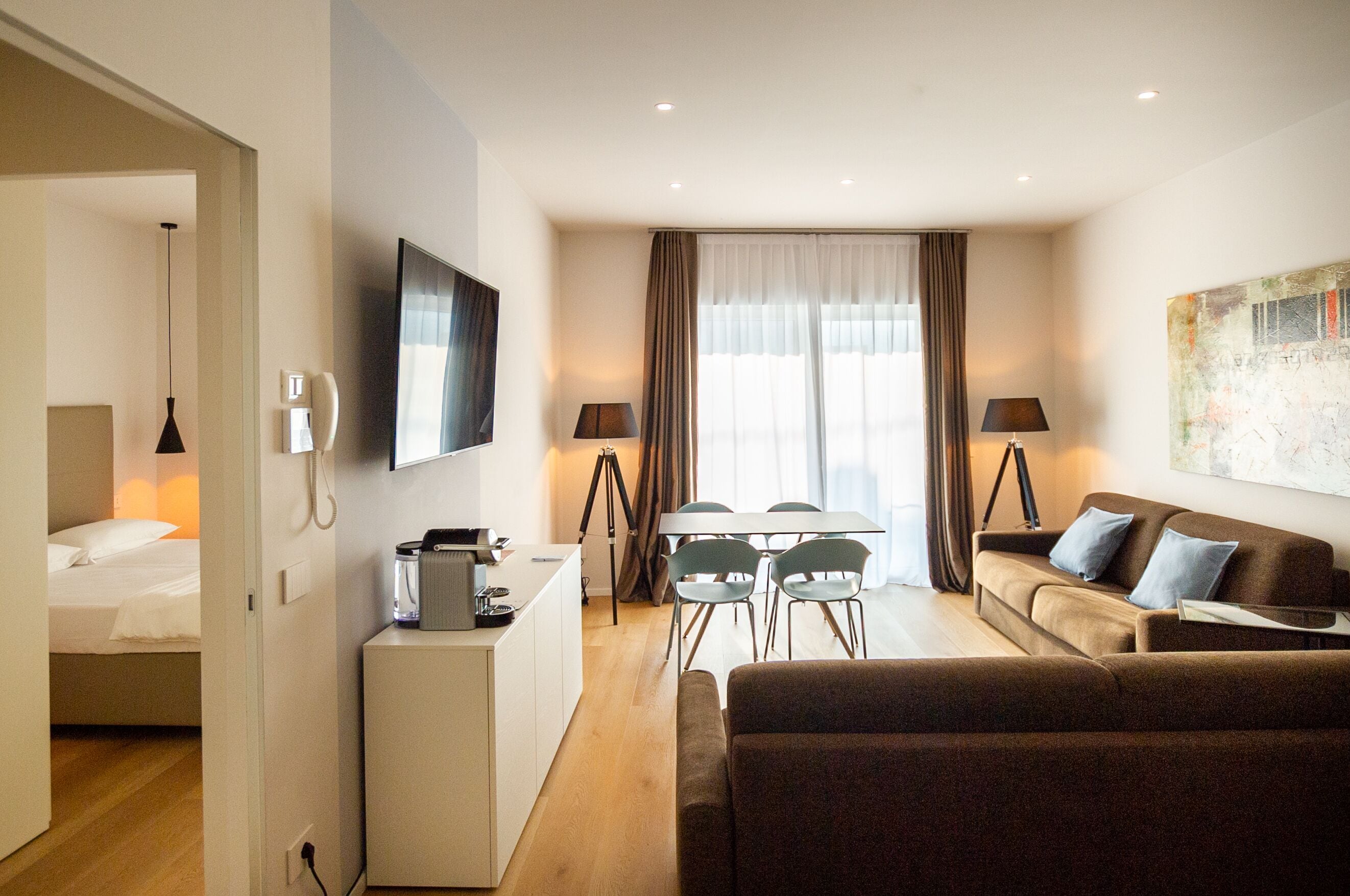 A compact hotel room with a bed, a desk with a computer, a dining table with chairs, a sofa, and a TV mounted on the wall.