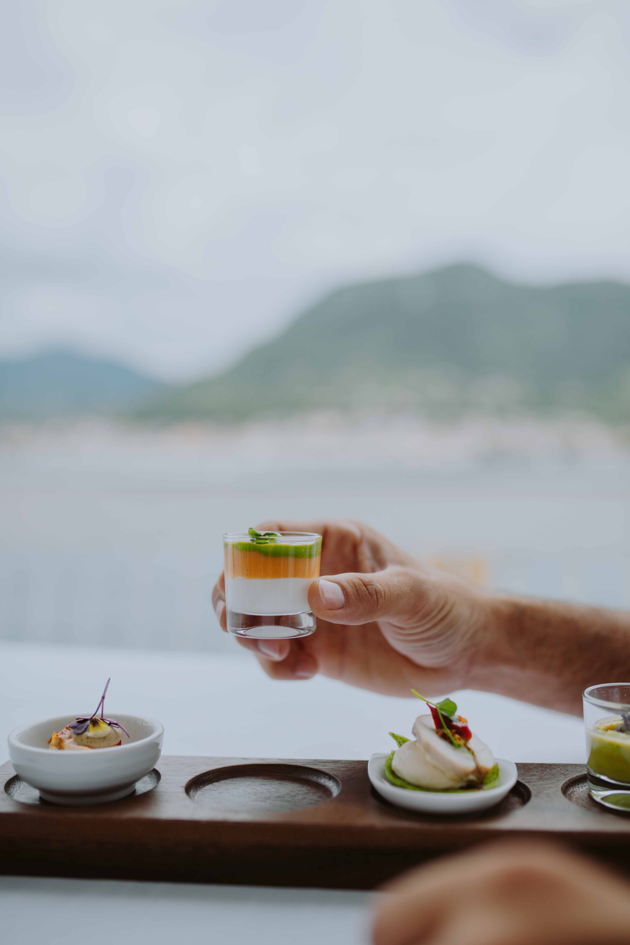Una mano sosteniendo un vaso con una bebida en capas, con una vista panorámica de montañas al fondo.