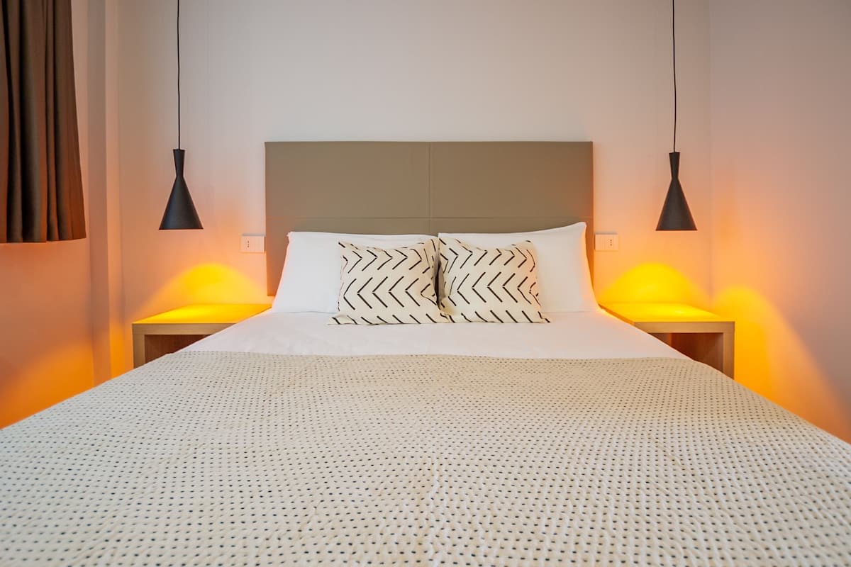 A neatly made bed with a patterned pillow, two bedside lamps, and a plain wall.