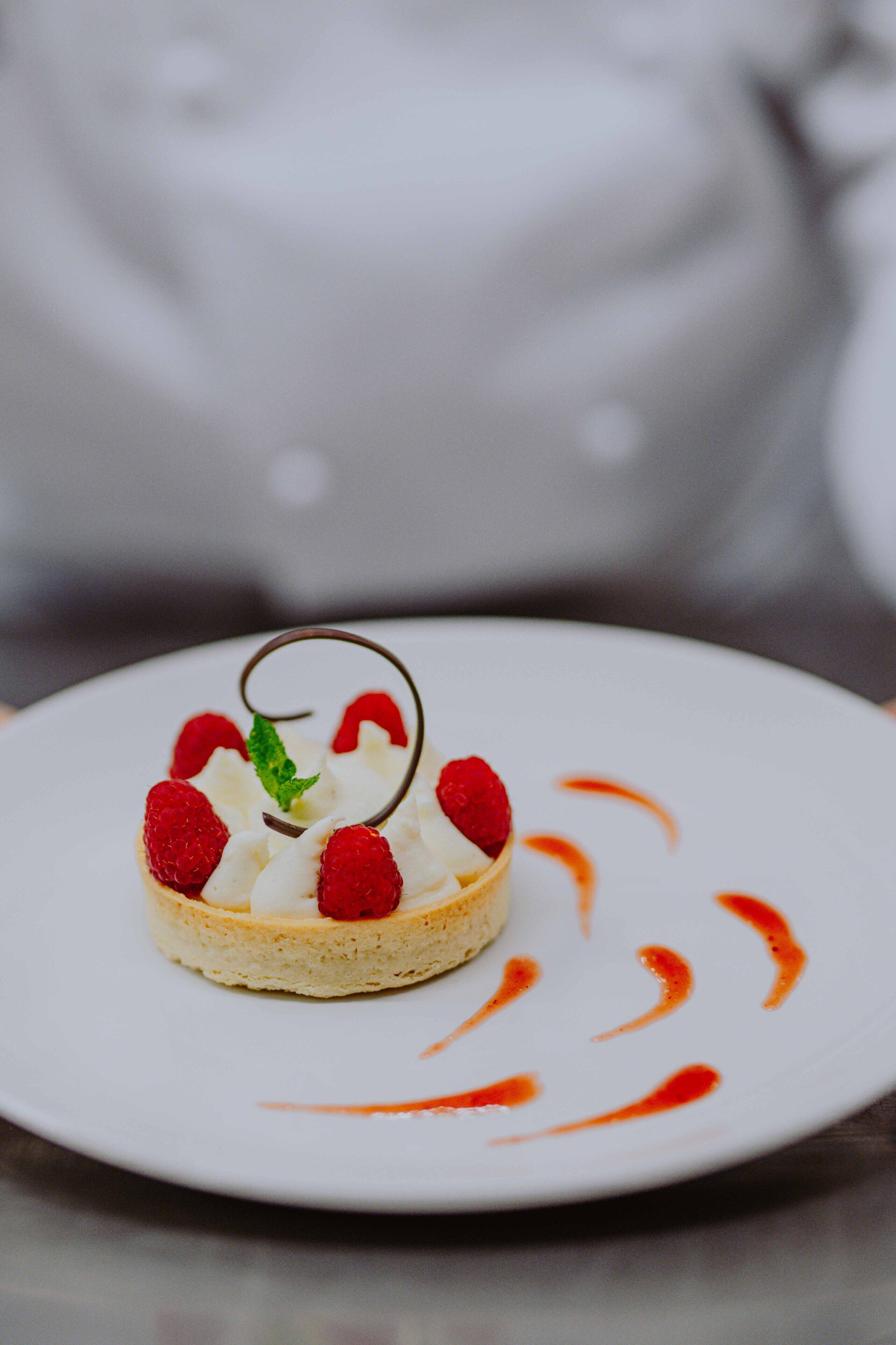 Un dessert su un piatto bianco, decorato con un filo di cioccolato, lamponi e una foglia di menta.