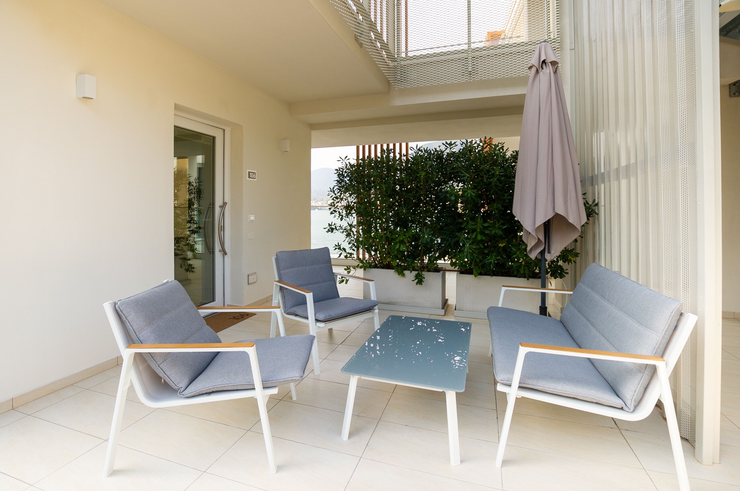 A modern outdoor seating area with white and gray chairs and a small table, surrounded by a white wall and a large window overlooking a garden.