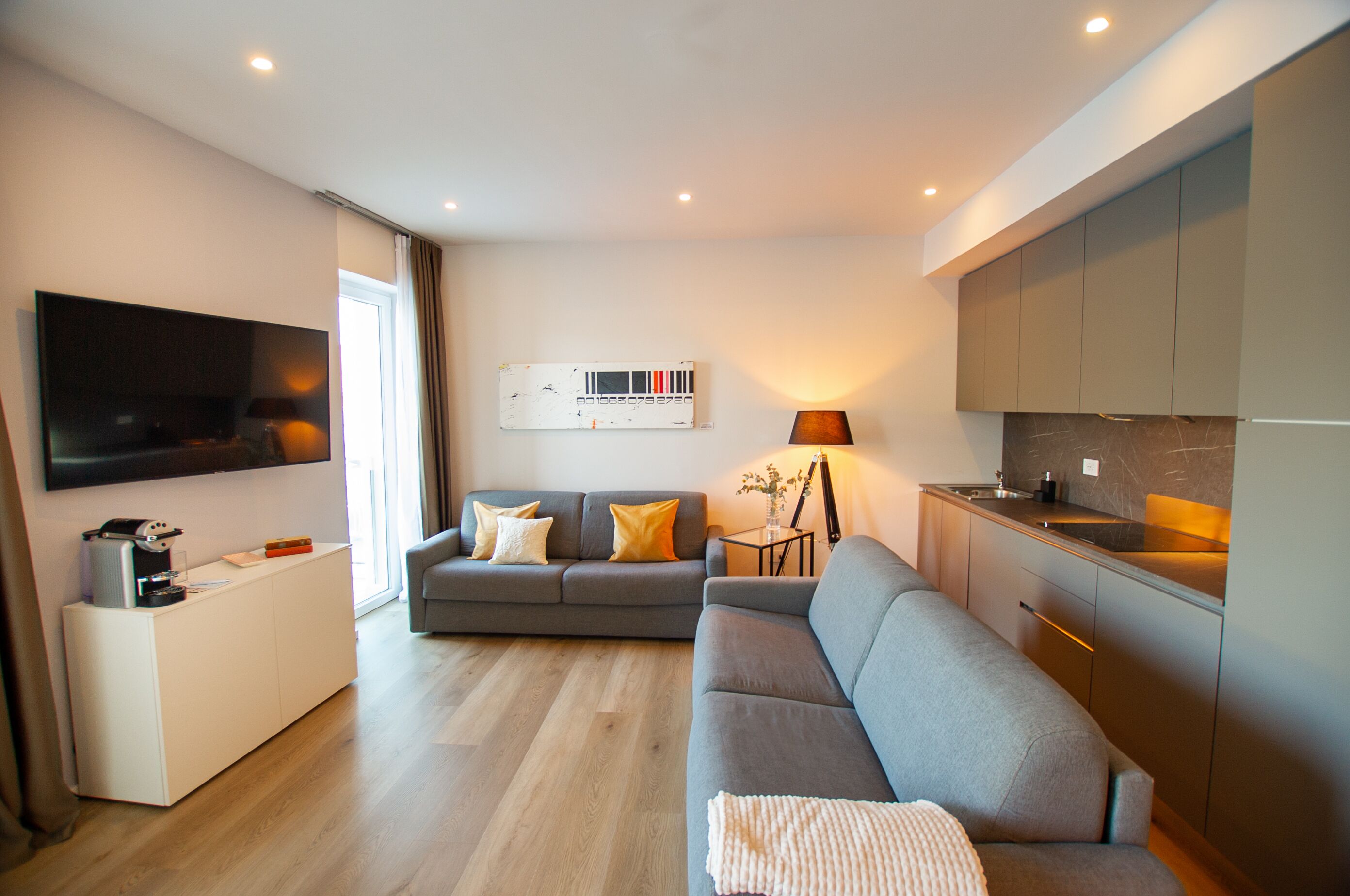 A modern living room with a grey sofa, a flat-screen TV mounted on the wall, and a small side table with a lamp.