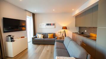 A modern living room with a grey sofa, a flat-screen TV mounted on the wall, and a small side table with a lamp.