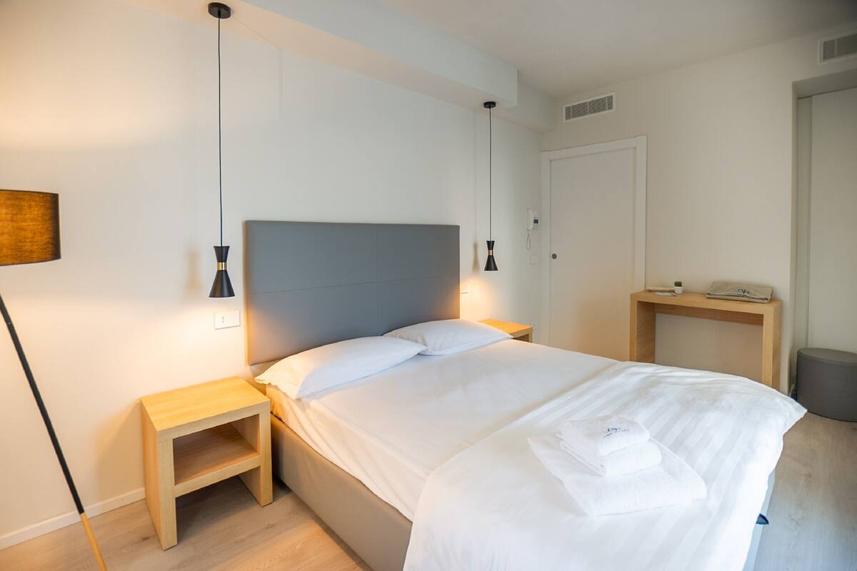 A neatly made bed with white linens and a grey headboard, two bedside tables, a lamp, and a small wooden nightstand.