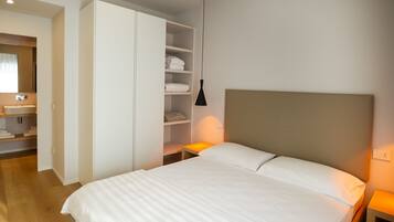 Habitació d'hotel amb llit, armari, taulella de nit amb làmpada i bany amb rentapla i mirall.