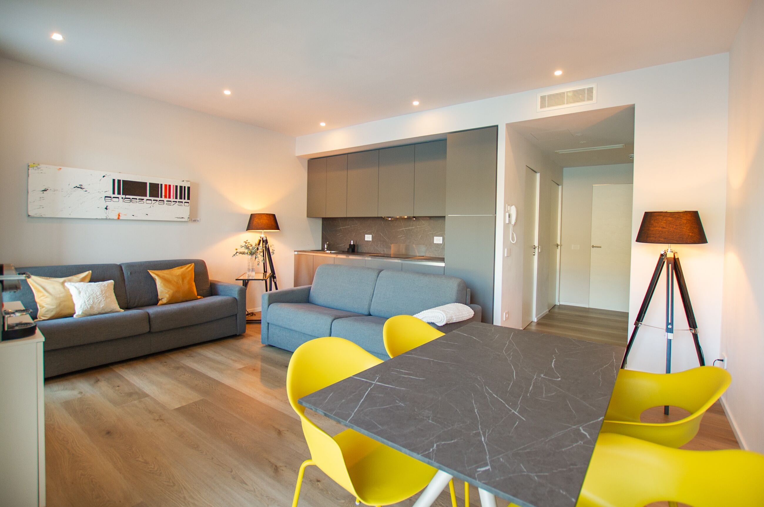 Een moderne woonkamer met een grijze bank, gele stoelen, een marmeren tafel en een keuken op de achtergrond.
