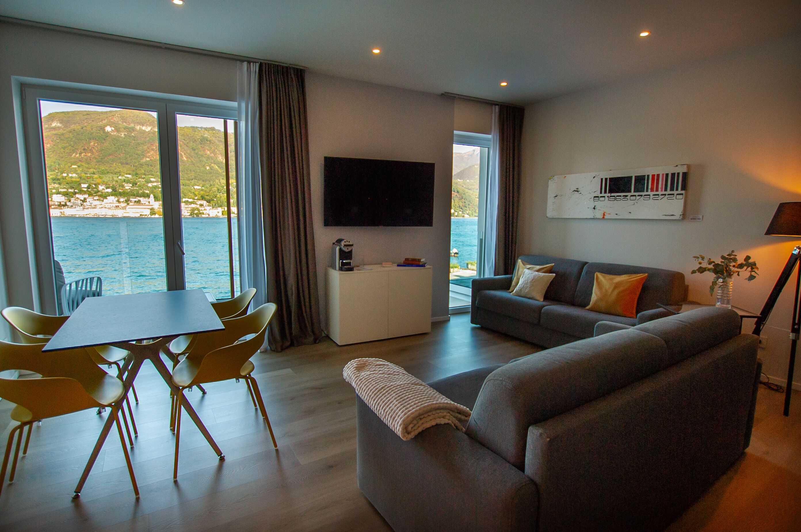 A modern living room with a sofa, a dining table with chairs, a flat-screen TV, and a view of the sea.