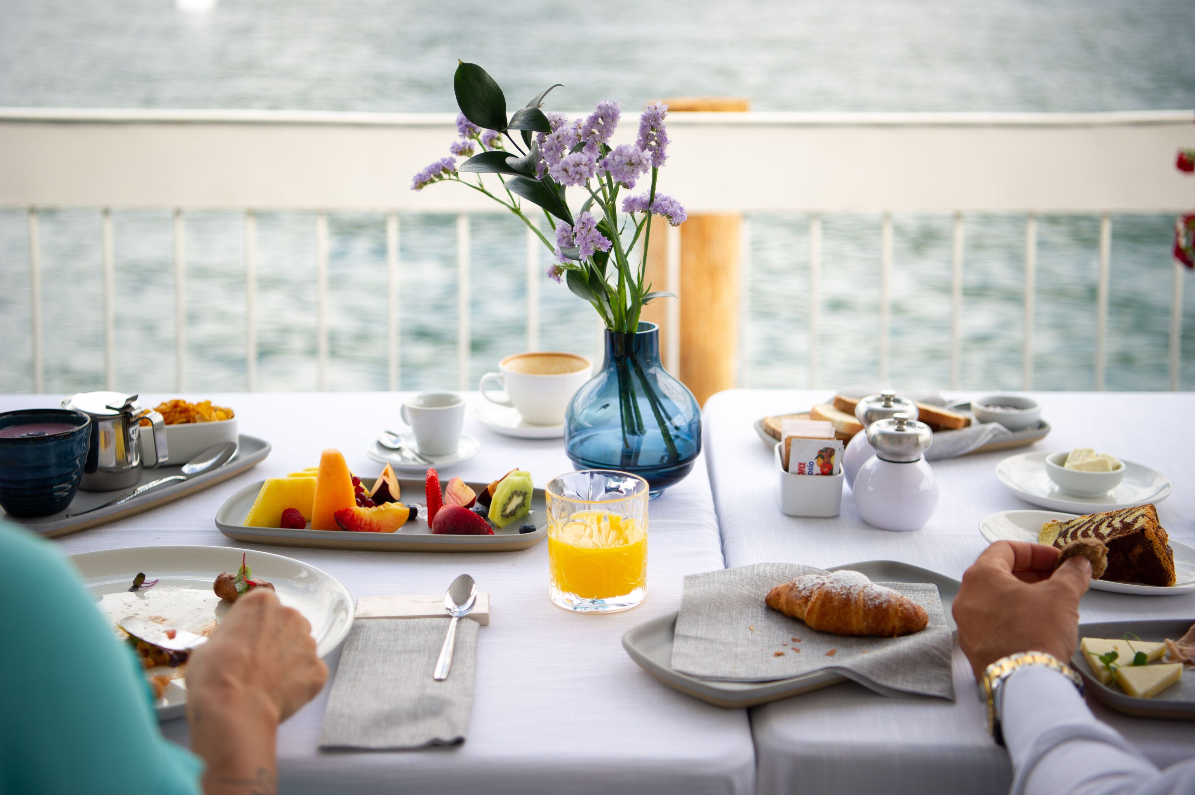 A breakfast table with various dishes, a vase with flowers, and a view of the sea.