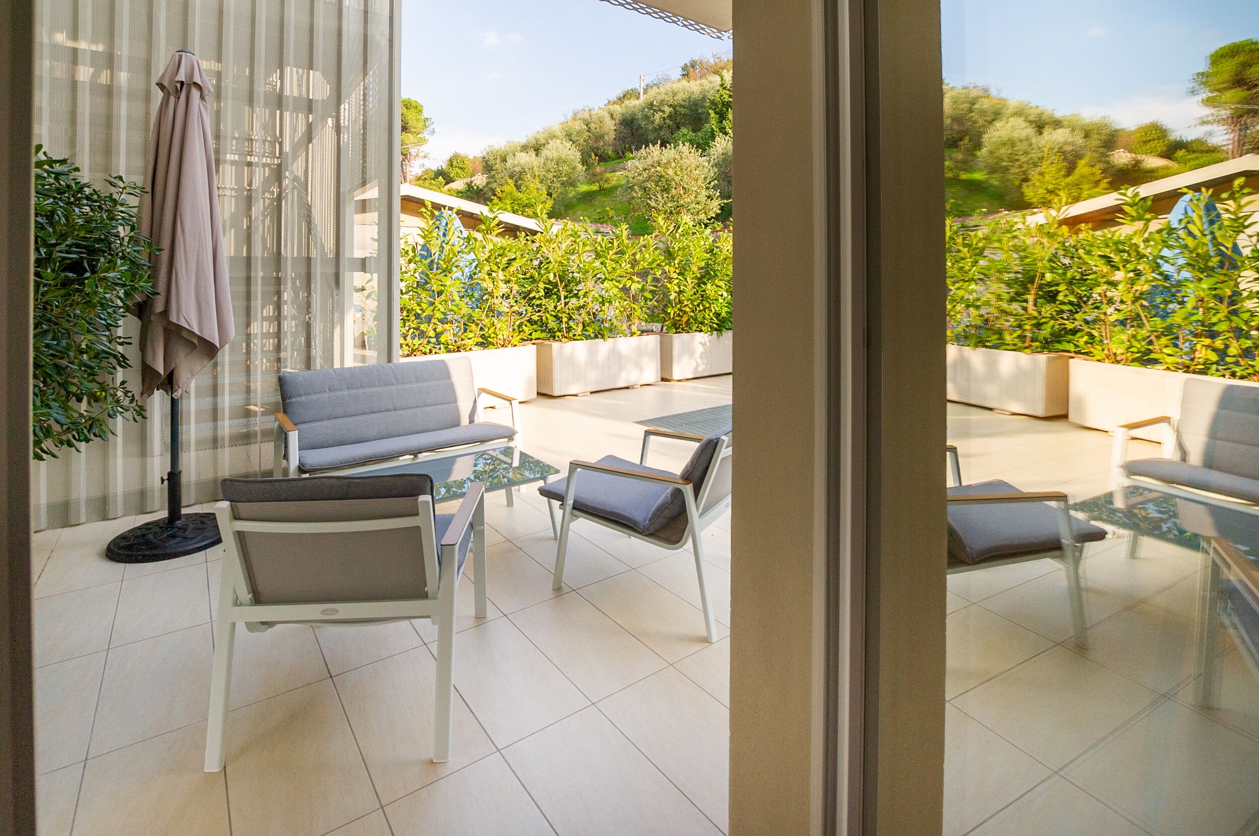 Une terrasse ensoleillée, agrémentée d’un canapé, de chaises et d’une petite table, donnant sur un jardin où poussent différentes plantes, avec une colline en arrière-plan.