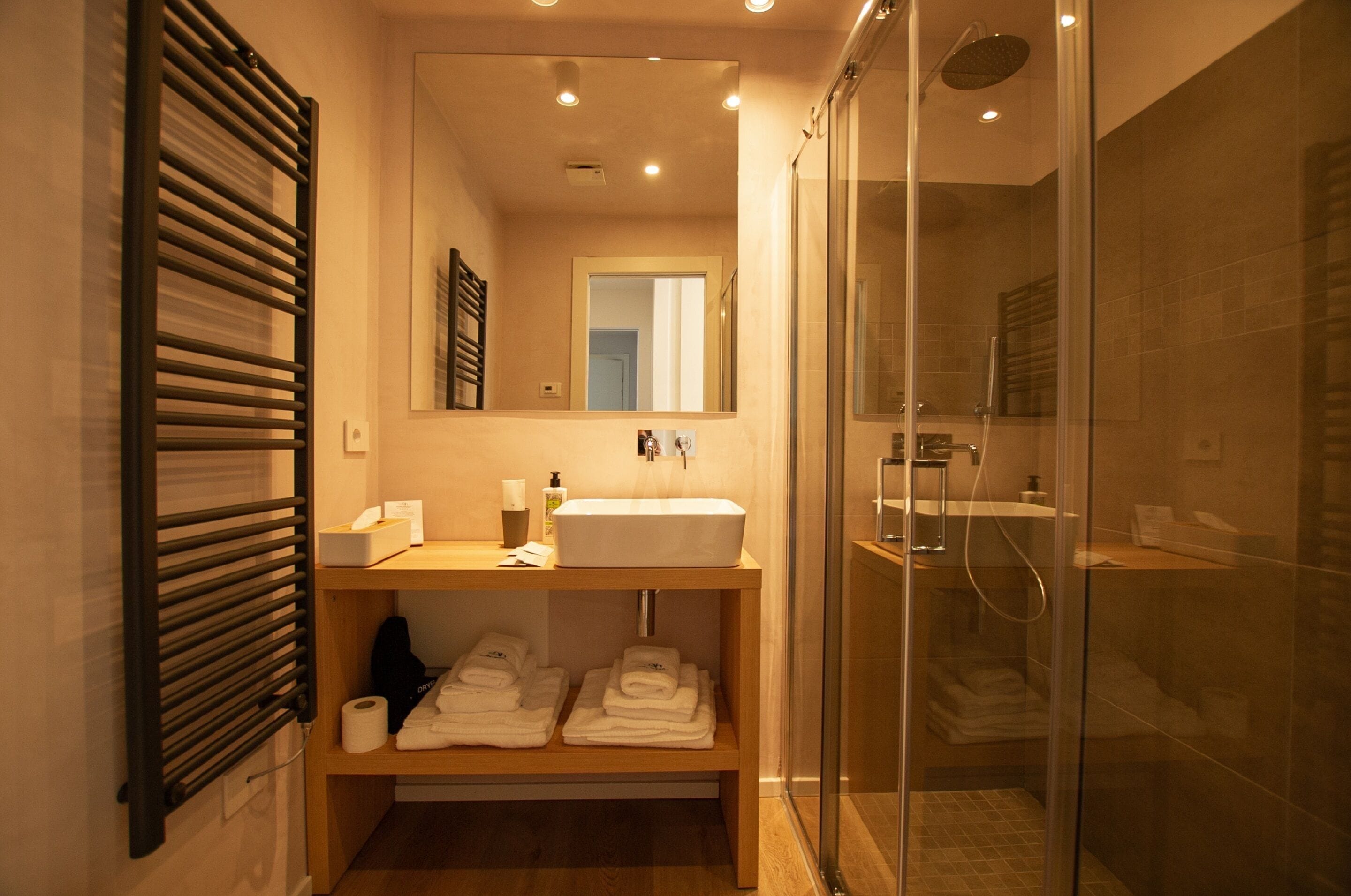 Un bagno moderno con doccia a vetro, un mobile bagno in legno con lavandino e asciugamani ordinatamente riposti su uno scaffale.