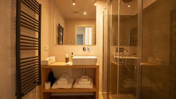 Un bagno moderno con doccia a vetro, un mobile bagno in legno con lavandino e asciugamani ordinatamente riposti su uno scaffale.