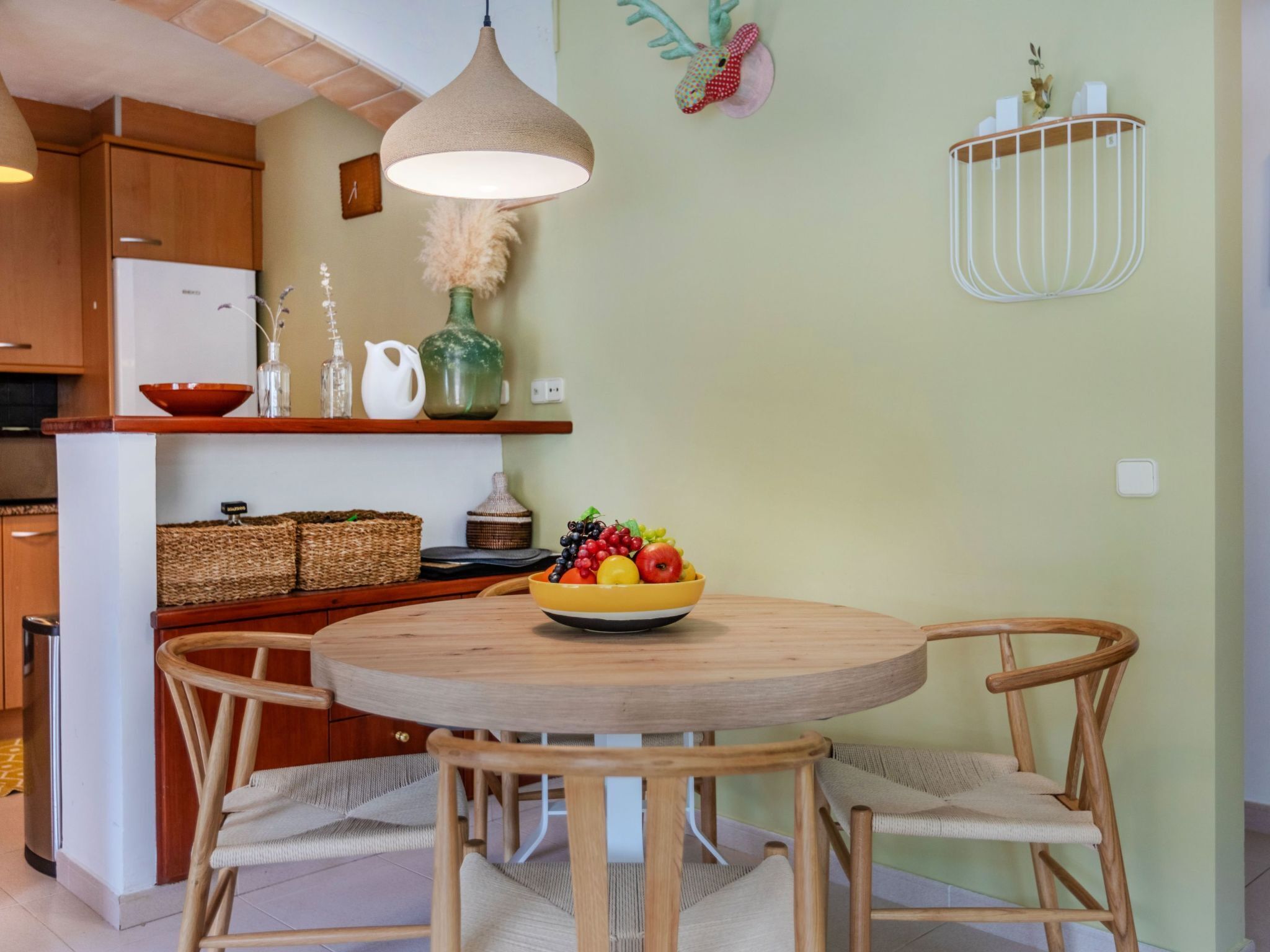 Een keuken met een ronde houten tafel, rieten stoelen, een hanglamp en een fruitschaal op de tafel.