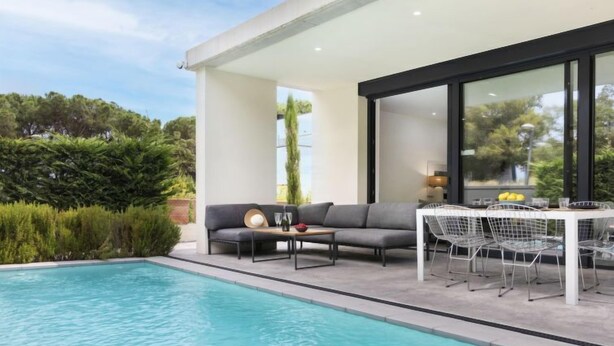 A modern outdoor pool area with a sofa and dining table, surrounded by greenery. - Vacation home Alyssa by Interhome (St Feliu de Guíxols)