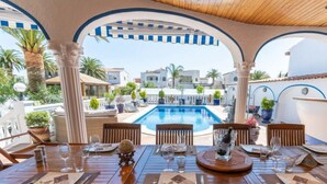 Un'area pranzo all'aperto con tavoli e sedie in legno, una piscina sullo sfondo e vista su edifici bianchi e palme.