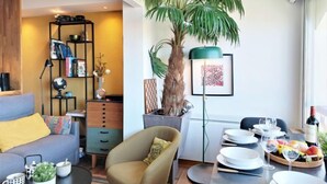 Ein modernes Wohnzimmer mit Sofa, Sessel, Couchtisch, Bücherregal und einem Essbereich mit Tellern und Gläsern.