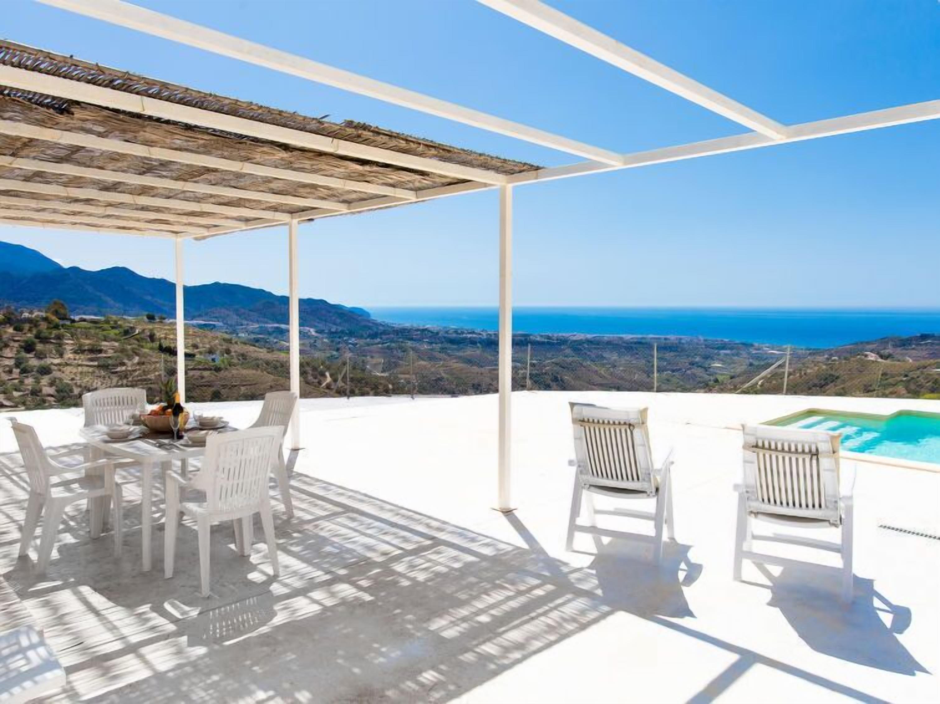 Un espace extérieur aménagé avec des sièges, protégé par un toit, comprenant des chaises blanches et une table, d’où l’on peut admirer un paysage montagneux et la mer.