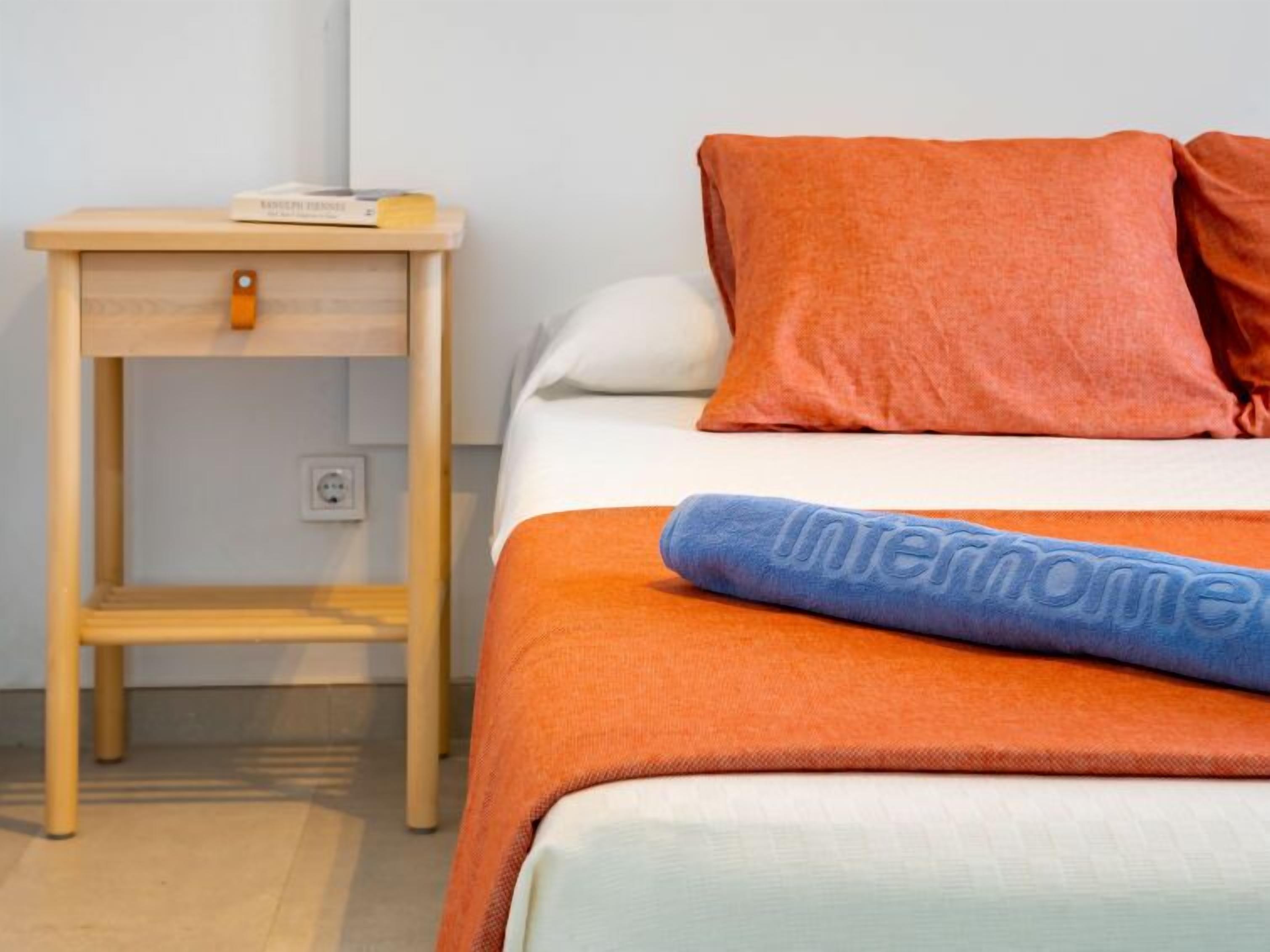 A neatly made bed with an orange blanket and pillows, a small wooden nightstand with a book and a lamp, and a power outlet on the wall.