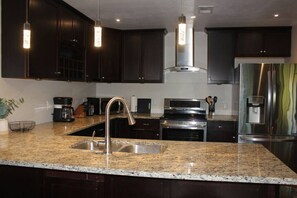 A modern kitchen with dark cabinetry, granite countertops, stainless steel appliances, and pendant lighting.