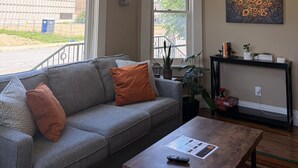 A living room with a grey sofa, a coffee table, a painting, and a ceiling light fixture. - 131 Cedar 2 · Chic 2BR Urban Retreat | Downtown Kalamazoo (Kalamazoo)